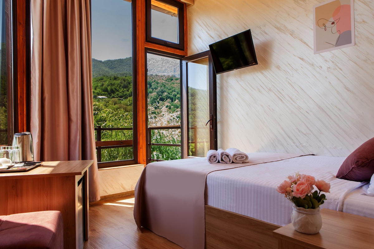 A bedroom with a bed, a desk, a chair, a TV, and a window with a view of greenery.
