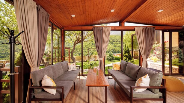 A spacious room with two grey sofas, a wooden table, and large windows offering a view of a lush garden. - Dilivita Rest House (Gosh)