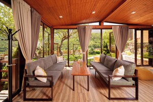 Una habitación amplia con dos sofás grises, una mesa de madera y grandes ventanales que dan a un jardín exuberante.