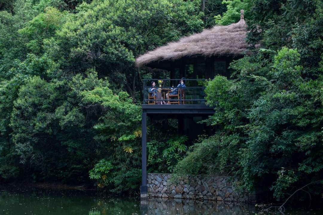 A thatched-roof structure with a stone base, surrounded by dense greenery and a body of water.