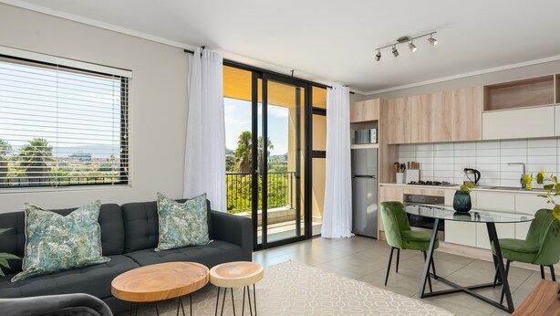 A modern living room with a sofa, two round wooden tables, and a dining area with a glass table and green chairs. - Waters Edge 109 (Cape Town)