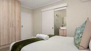 A bedroom with a bed, a green throw blanket, a rolled towel, a wooden wardrobe, and a bathroom with a glass shower and a sink. - Waters Edge 109 (Cape Town)