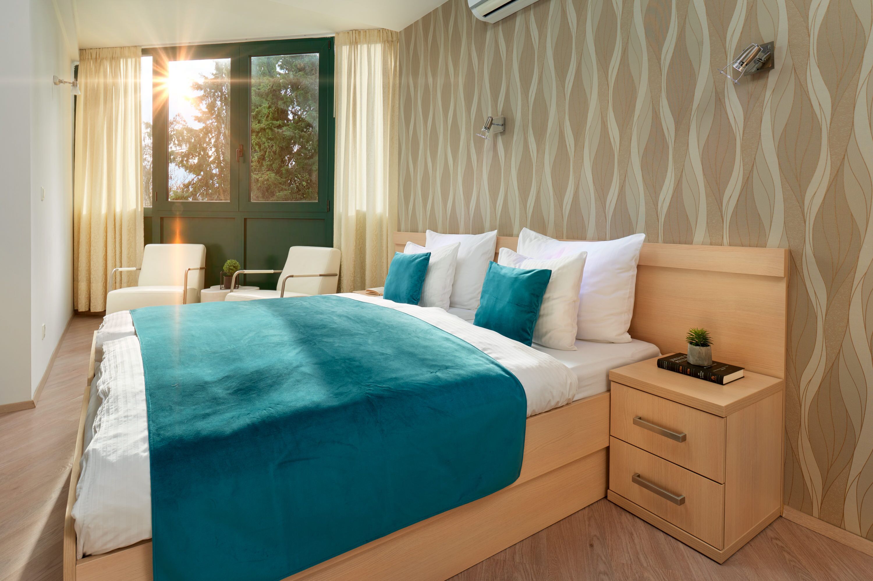 A modern hotel room with a large bed, a bedside table with a lamp, and a view of trees through the window.