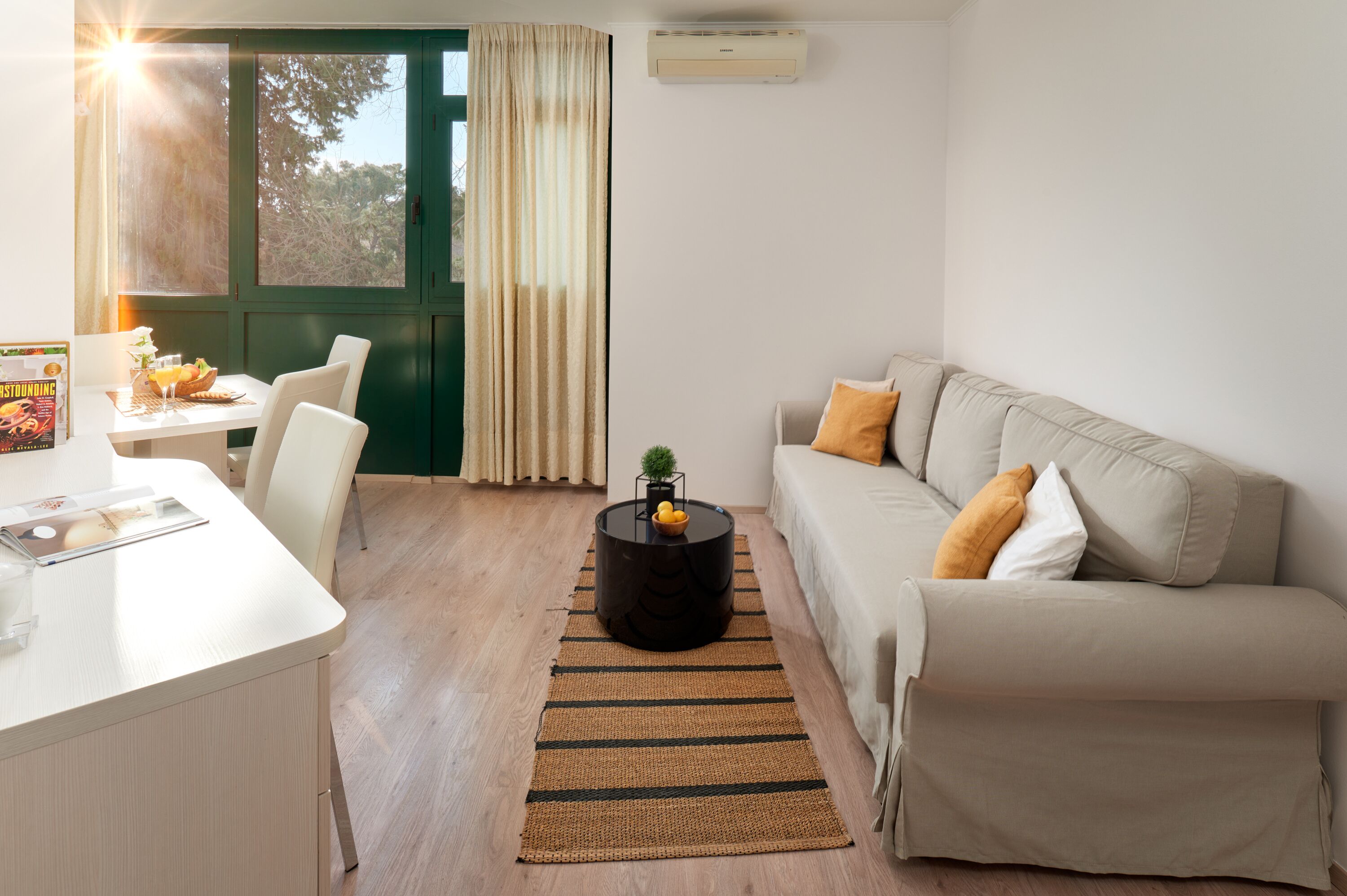 A modern living room with a white sofa, a small round table with a bowl of fruit, a white desk with a magazine, and a large window with curtains.