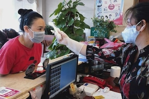 Una persona con una camisa roja frente a un mostrador con un monitor de computadora, una planta y varios objetos.