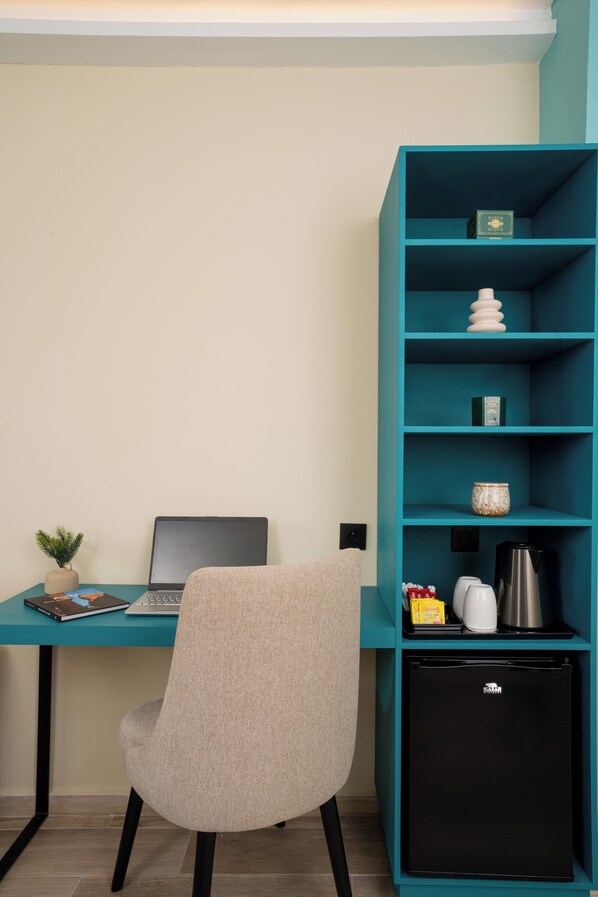 Un escritorio color turquesa con una computadora portátil, una planta pequeña y una silla. A la derecha, una estantería color turquesa con un refrigerador negro y varios objetos.