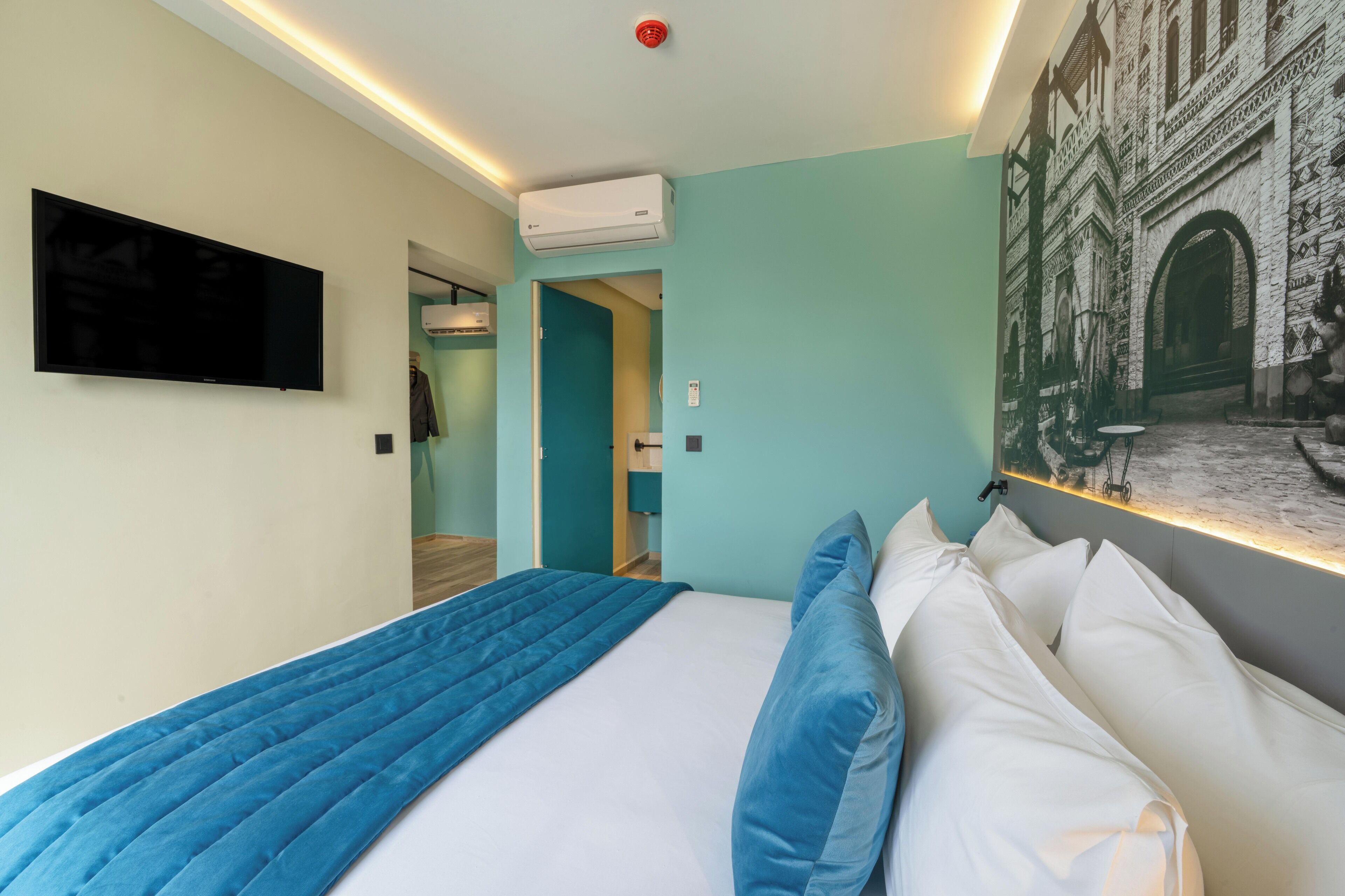 Ein modernes Hotelzimmer mit einem großen Bett, einem Flachbildfernseher, einer Klimaanlage und einem Wandbild, das ein historisches Gebäude zeigt.