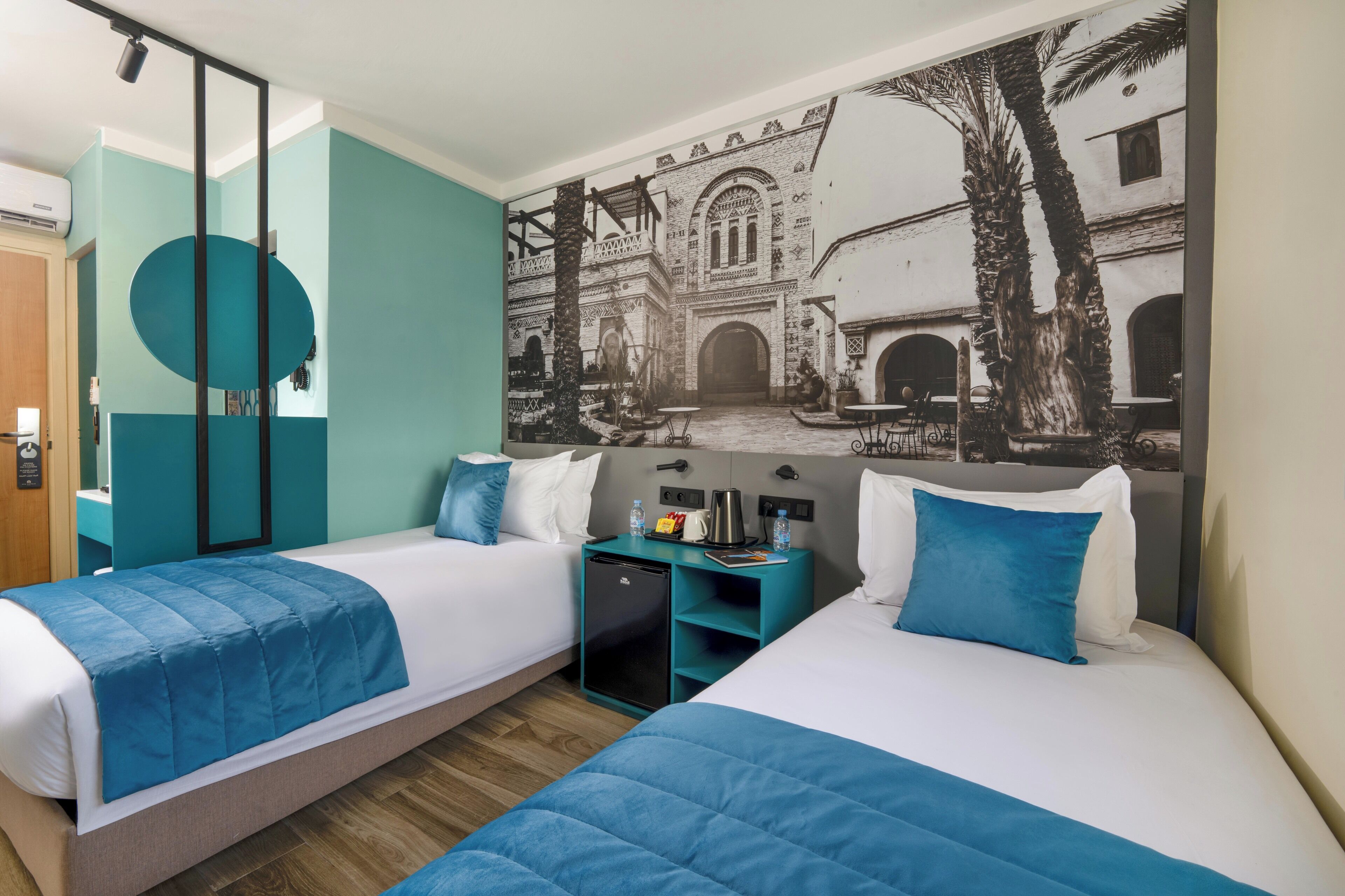 Hotelkamer met twee bedden, blauwgroene wanden, een muurschildering van een binnentuin en een klein bureau met koffiezetapparaat.