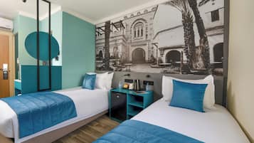 A hotel room with two beds, teal walls, a mural of a courtyard, and a small desk with a coffee maker.