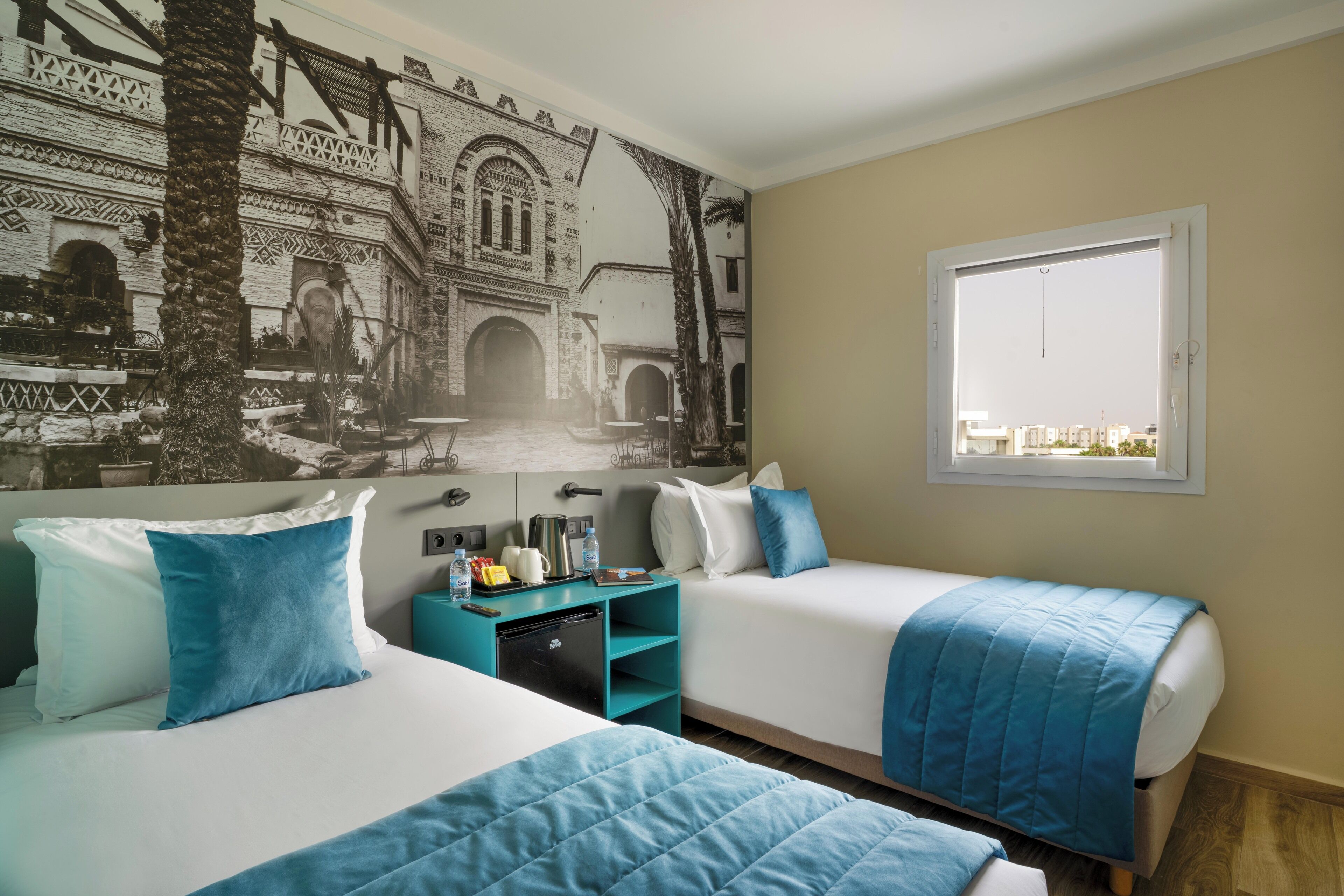 Habitación de hotel con dos camas, un mural de una estructura arquitectónica, una ventana con vistas y una pequeña mesita auxiliar azul verdosa con comodidades.
