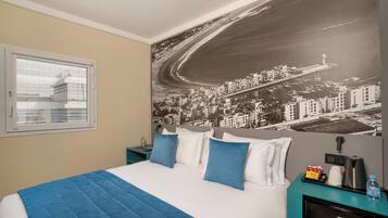A hotel room with a bed, a cityscape mural on the wall, a window, and a nightstand with a kettle and a bottle.