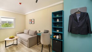 A modern living room with a sofa, a small table with fruit, a desk with a laptop, a teal bookshelf, and a coat hanging on the wall. - Mia Hotels Agadir (Agadir)