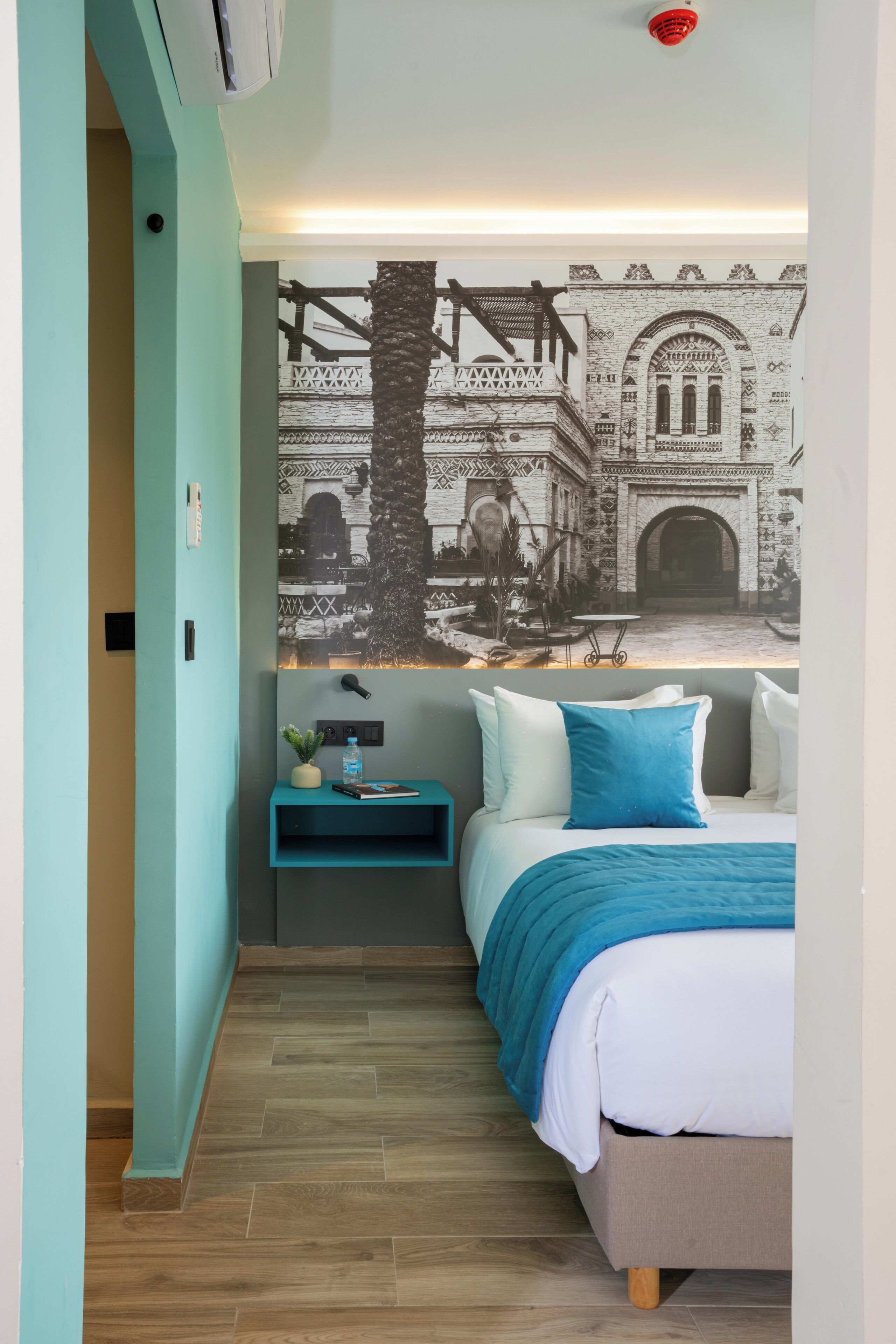 Un dormitorio con cama, mesita de noche, estante en la pared y un mural de un edificio.
