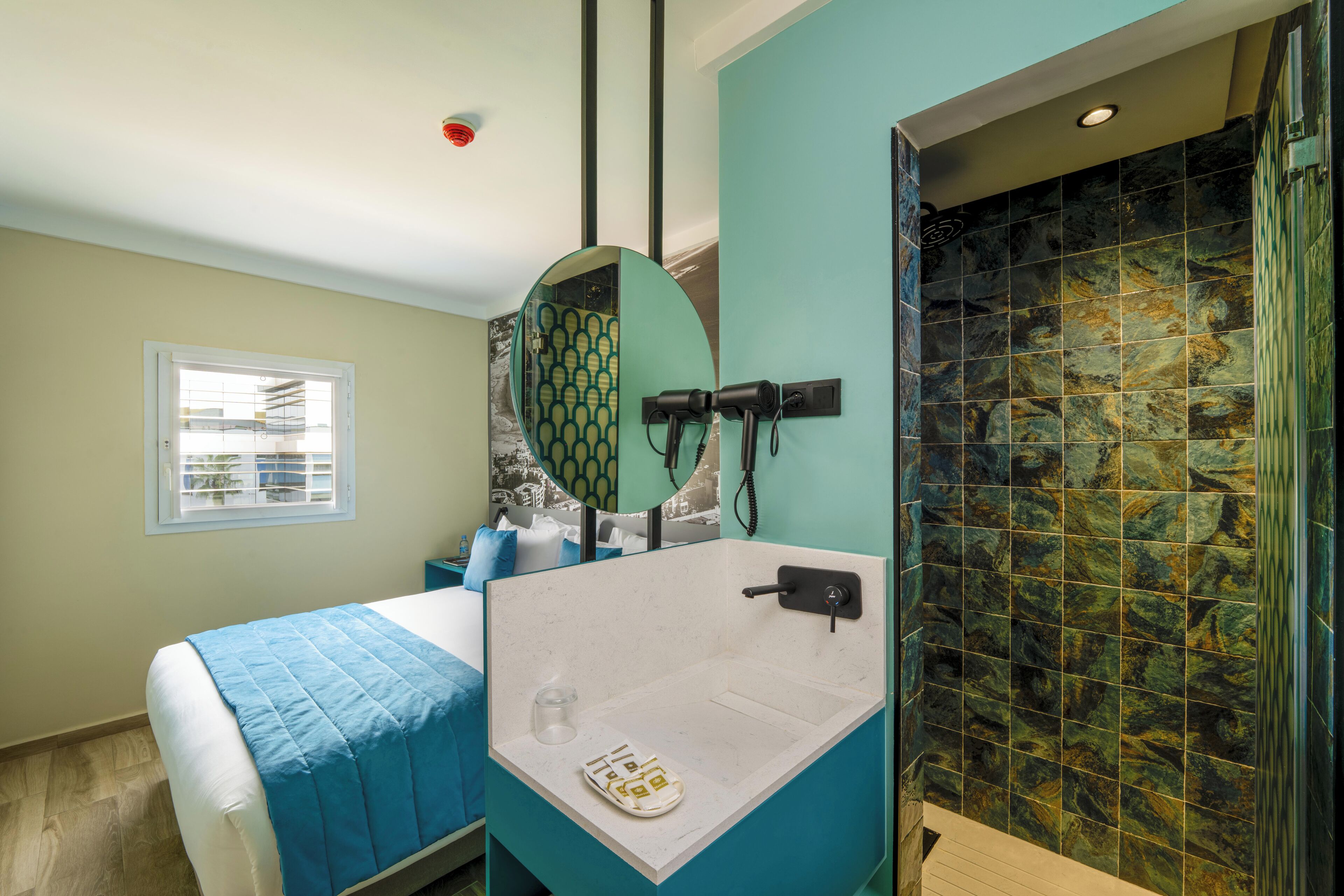 Una habitación de hotel con una cama, un baño con lavamanos y espejo, y una ventana con persianas.