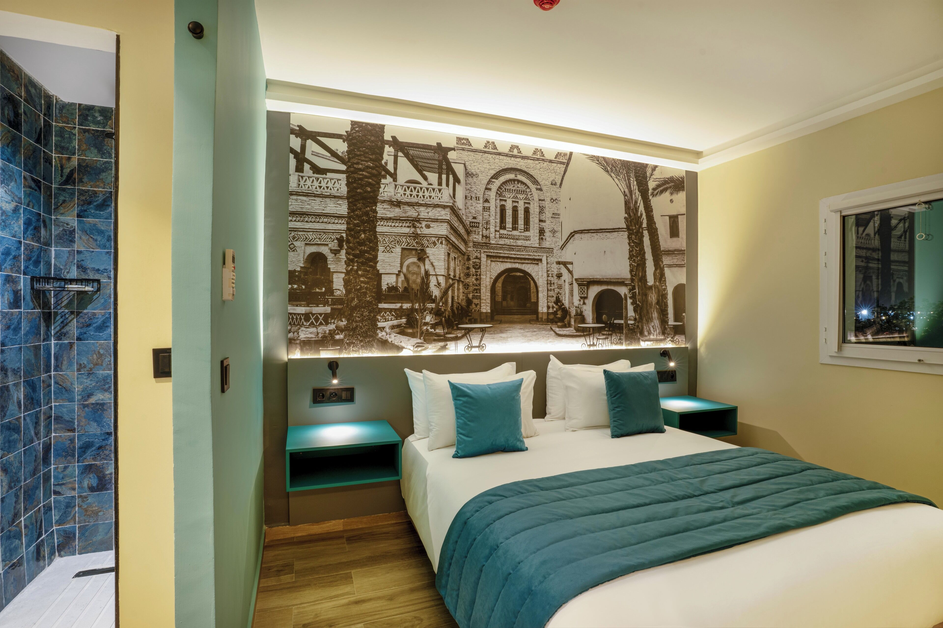 A hotel room with a large bed, a nightstand, a wall-mounted TV, and a bathroom with blue tiles.