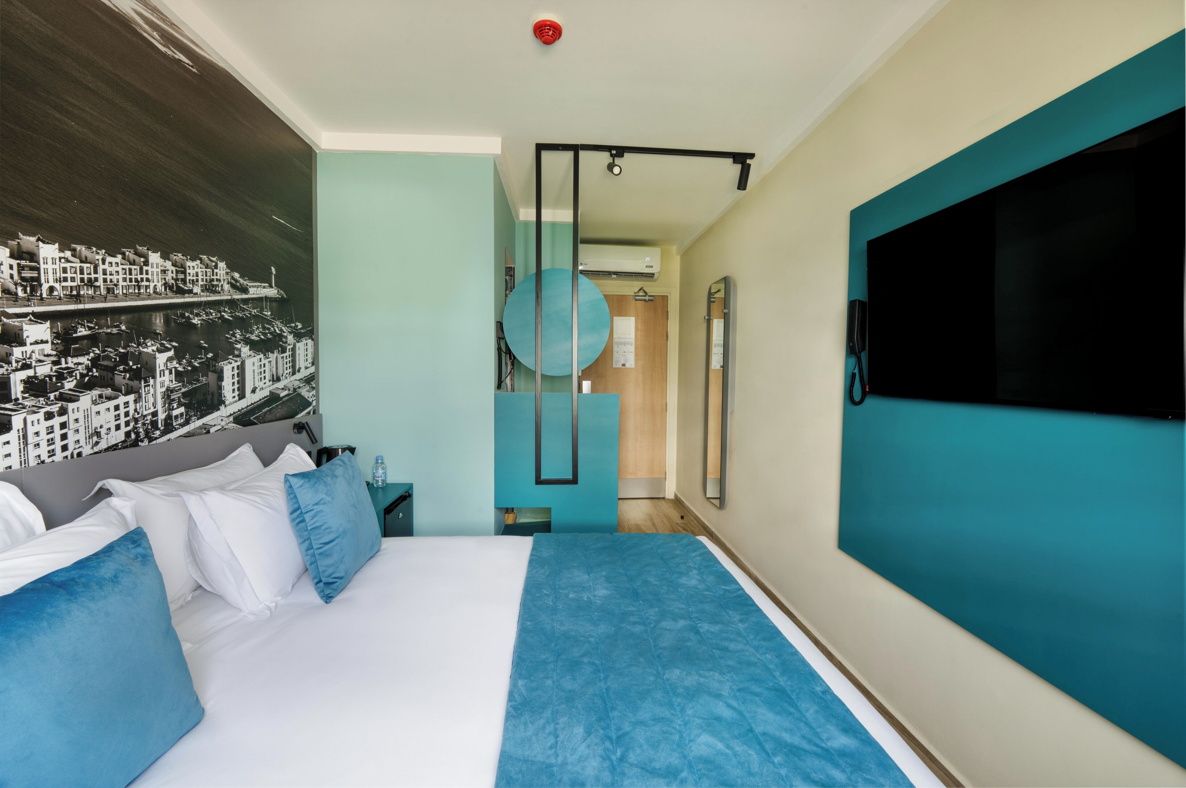 Habitación de hotel moderna con una cama, un televisor grande, un espejo circular fijado a la pared y un mural con vista a la ciudad.