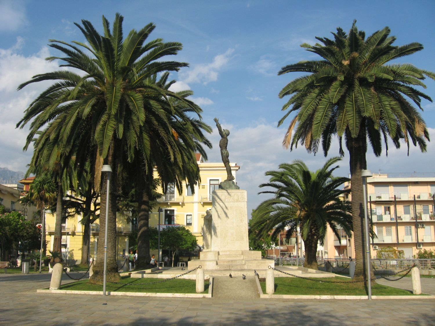 Une place publique bordée de palmiers, avec un monument et des bâtiments en arrière-plan.