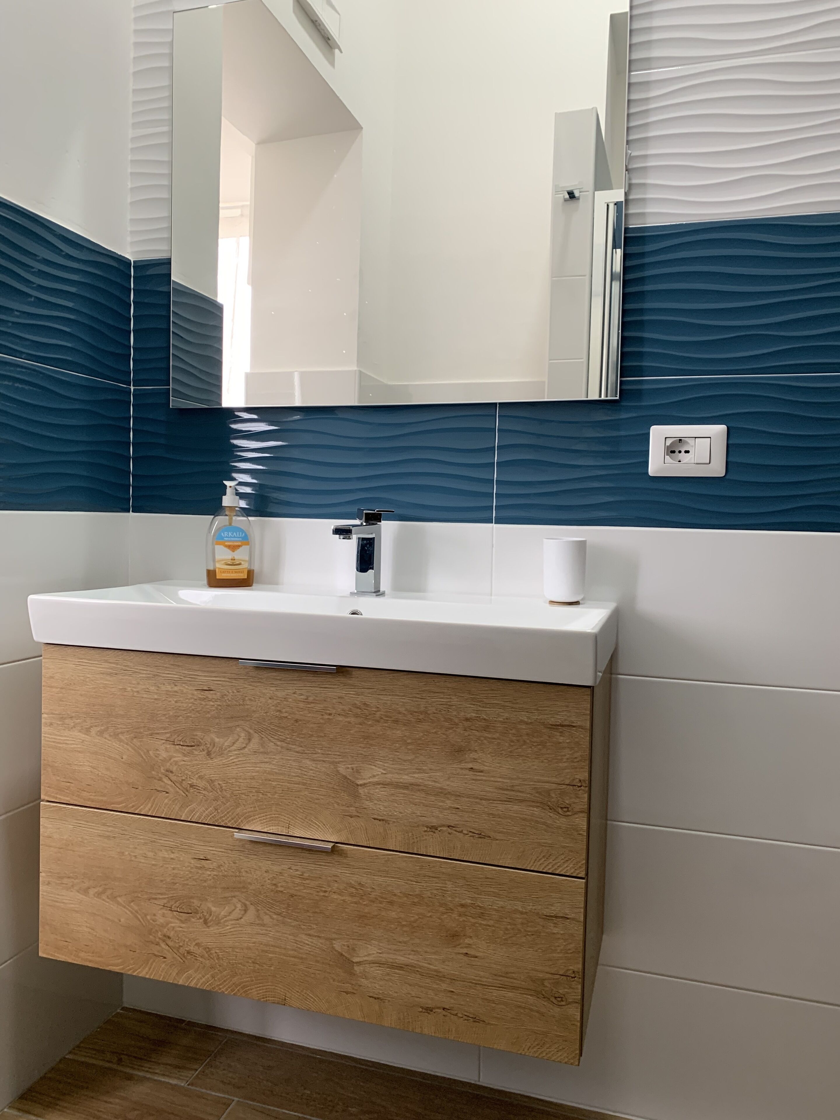 Une salle de bain avec un lavabo blanc, des tiroirs en bois, un miroir et des carreaux bleus à motifs de vagues.