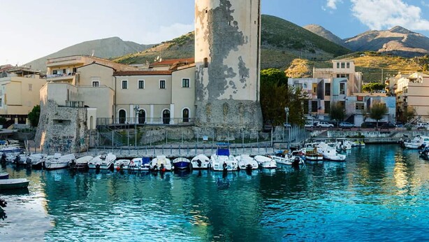 Een kustplaats met een opvallende toren, een jachthaven met boten, en gebouwen in mediterrane stijl.