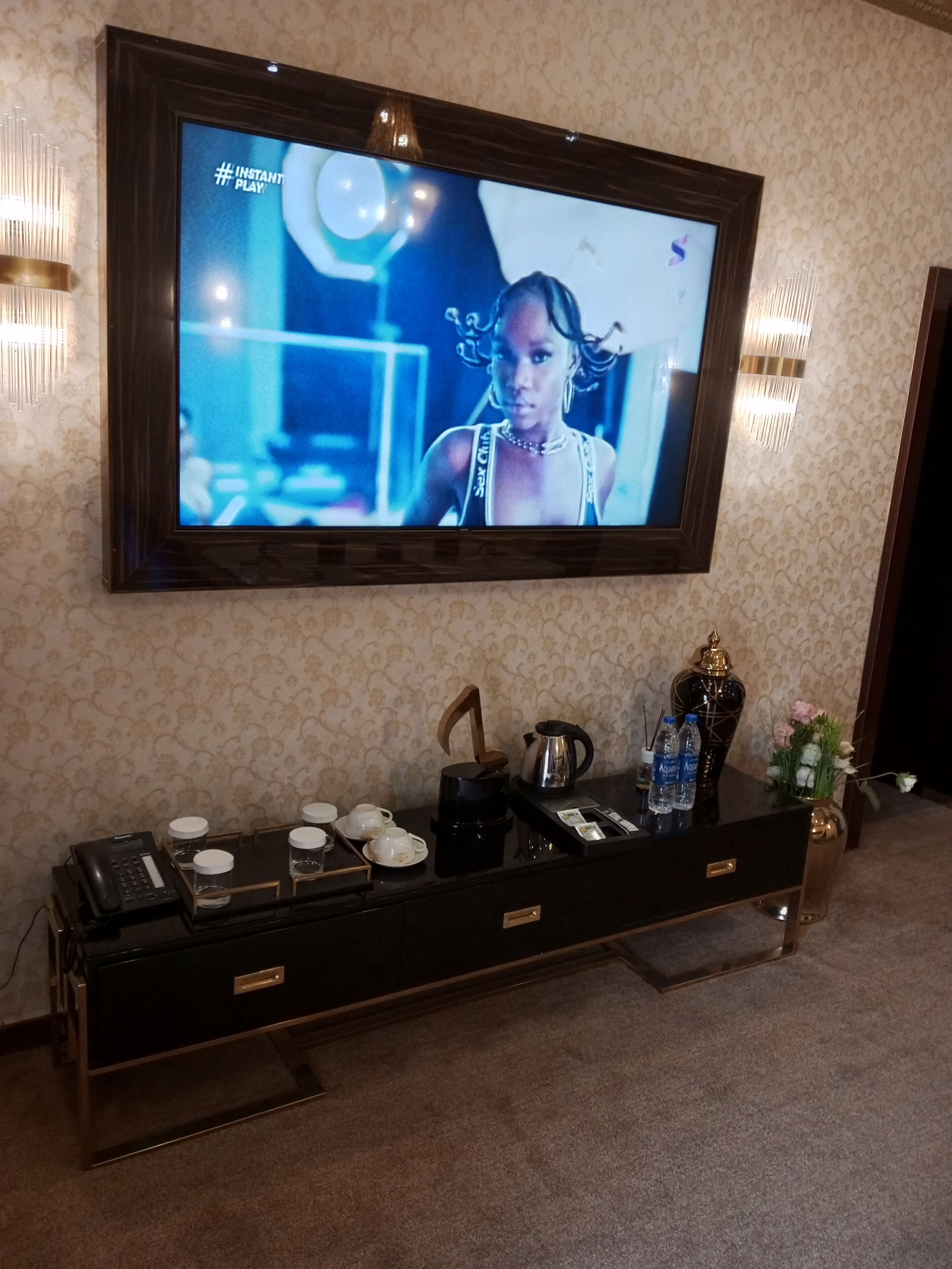 A television mounted on a wall displaying a show, a coffee maker, cups, and a vase with flowers on a console table.