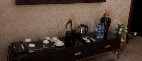 A television mounted on a wall displaying a show, a coffee maker, cups, and a vase with flowers on a console table.