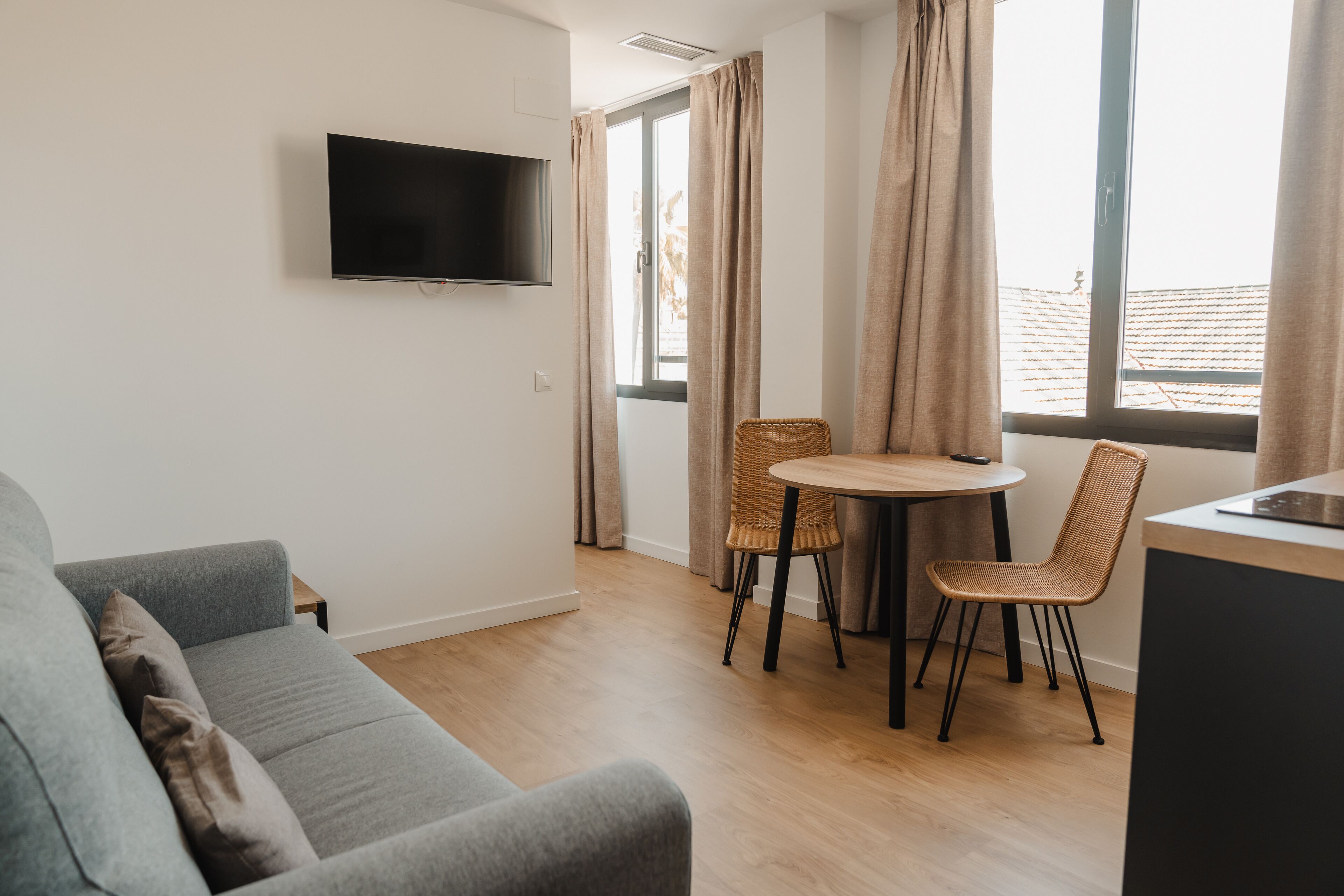 A modern living room with a grey sofa, a flat-screen TV mounted on the wall, a dining area with a round table and two wicker chairs, and large windows with curtains.