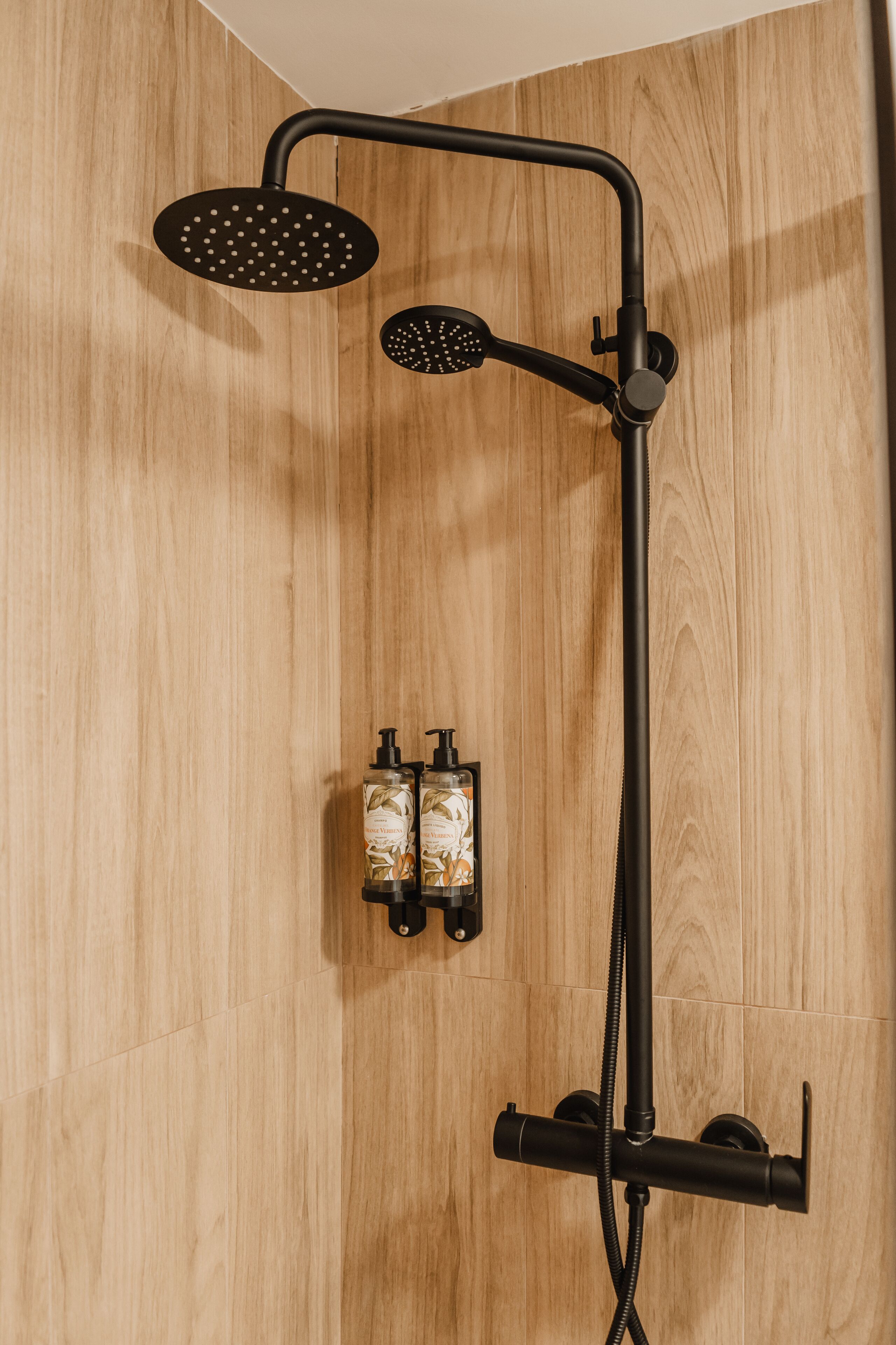 A shower with a rain showerhead and handheld showerhead, a handheld showerhead, and two bottles of shampoo and conditioner mounted on the wall.
