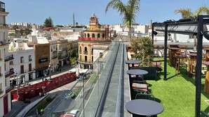 Terraza en la azotea con barandilla de cristal, mobiliario exterior y vistas a la ciudad, incluyendo edificios históricos y una palmera.