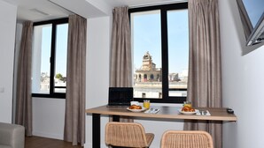Habitación moderna con una mesa de comedor, dos sillas de mimbre, una computadora portátil, alimentos para el desayuno y vistas de la ciudad a través de ventanas amplias.