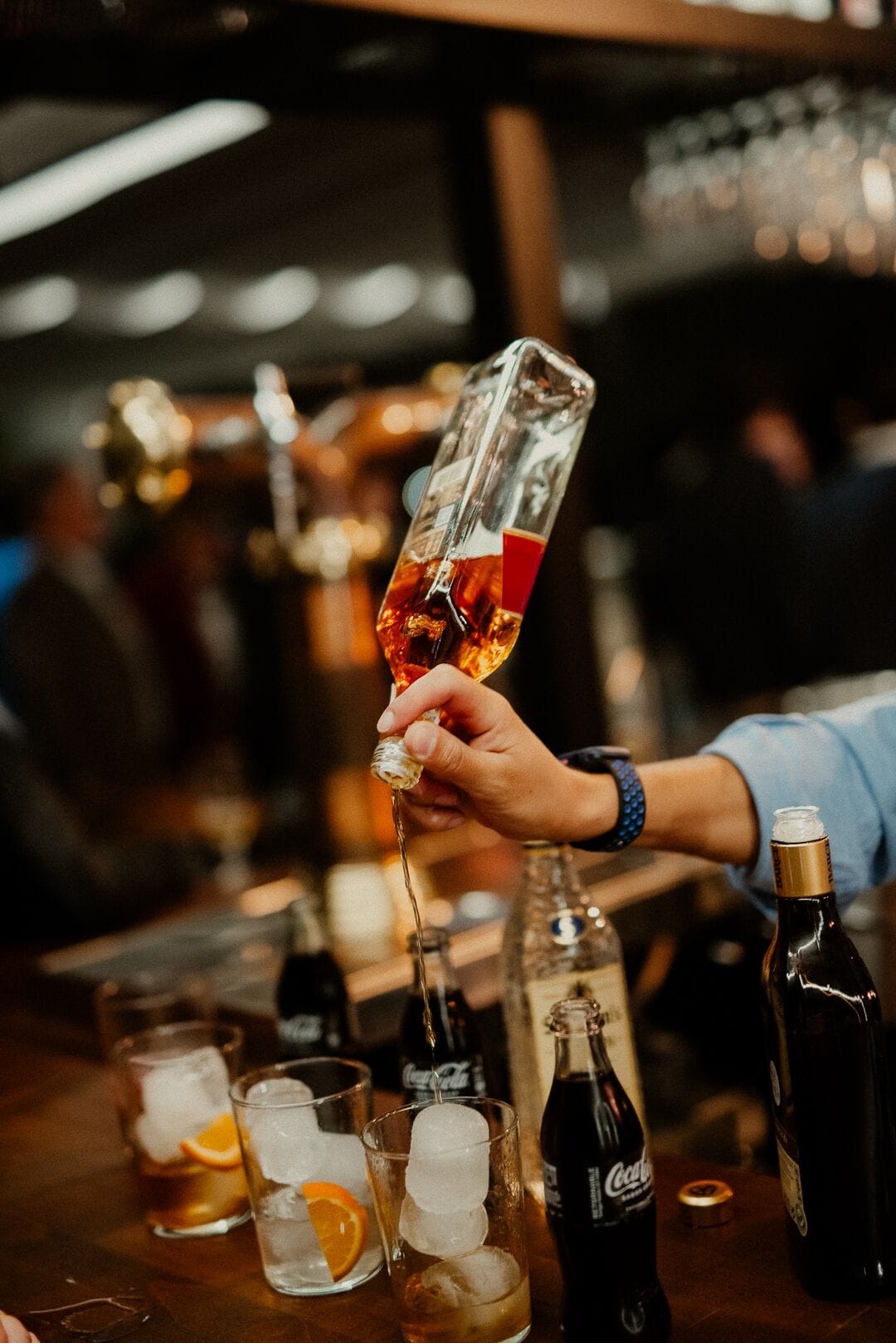 Een bartender schenkt een drankje in een glas met ijs en een oranje schijfje. Op de bar staan flessen Coca-Cola en andere dranken.