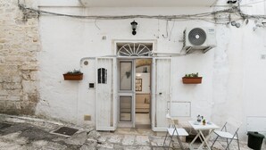Ein kleines, weißes Gebäude mit offener Tür zu einem Raum. Vor der Tür stehen ein Tisch und zwei Stühle, an der Wand steht ein Blumentopf und eine Klimaanlage ist an der Außenwand angebracht.