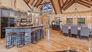 A spacious kitchen with a large island, bar stools, and a dining area with a high ceiling and wooden beams. - Indigo Falls ~ 8BR w/Golf Simulator, Indoor Cave Pool, Hot Tub, Game Room, Theater, & Views! (Sevierville)