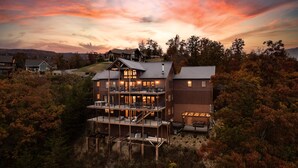 A multi-story building with a balcony and outdoor seating area, surrounded by trees and set against a sunset sky. - Indigo Falls ~ 8BR w/Golf Simulator, Indoor Cave Pool, Hot Tub, Game Room, Theater, & Views! (Sevierville)