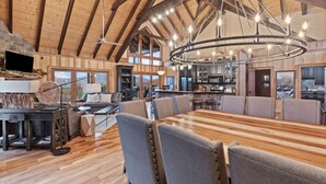 A spacious dining area with a long wooden table and several upholstered chairs, a stone fireplace, and a large chandelier. - Indigo Falls ~ 8BR w/Golf Simulator, Indoor Cave Pool, Hot Tub, Game Room, Theater, & Views! (Sevierville)