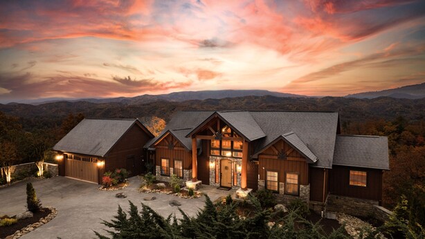 A large, multi-story log cabin with a stone foundation, multiple chimneys, and a driveway leading to a gated entrance. - Indigo Falls ~ 8BR w/Golf Simulator, Indoor Cave Pool, Hot Tub, Game Room, Theater, & Views! (Sevierville)