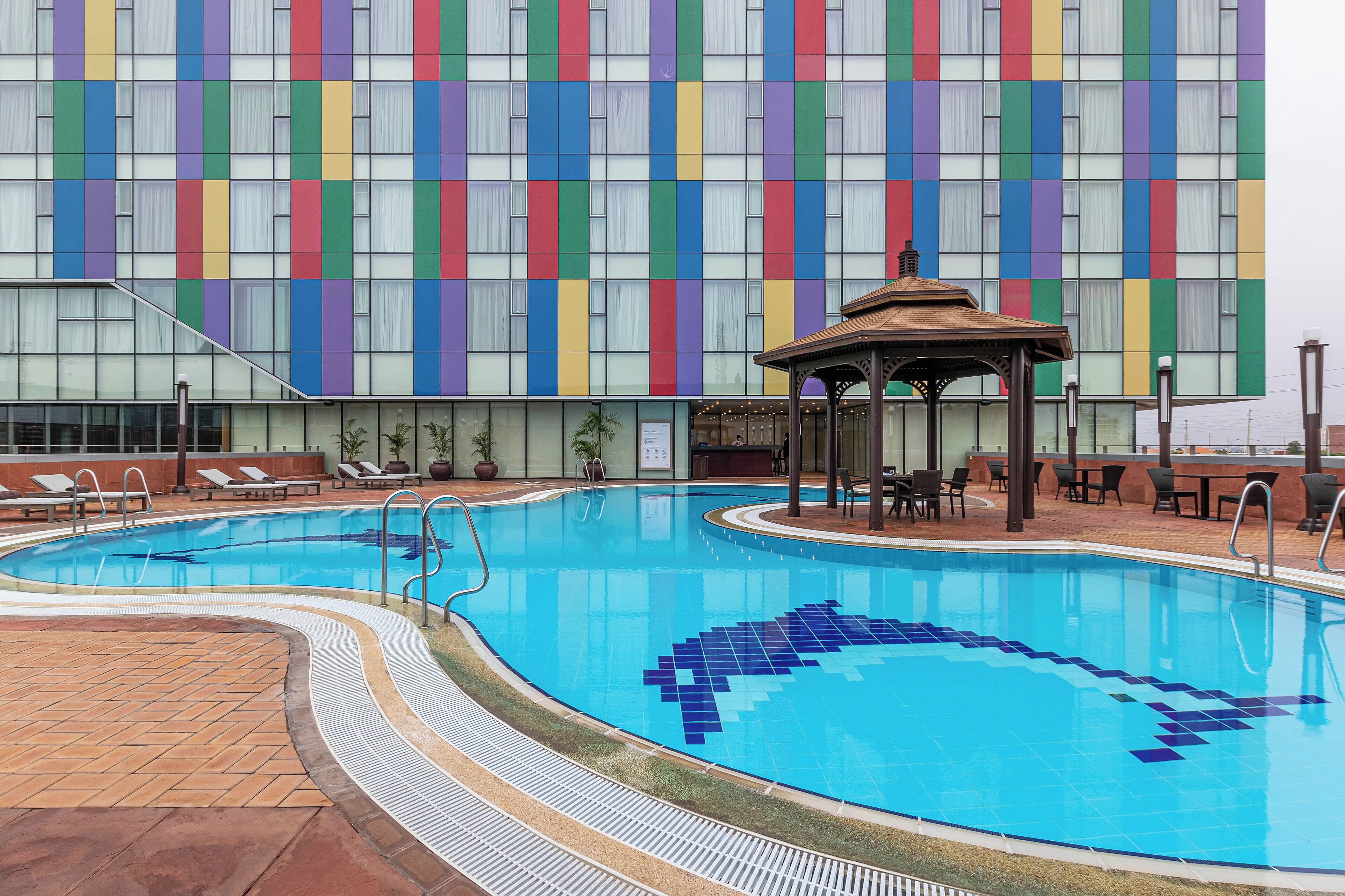 A swimming pool with a tiled design, a gazebo, lounge chairs, and a building with a colorful facade.