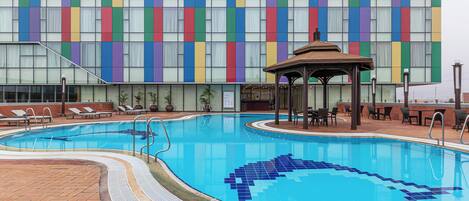 A swimming pool with a tiled design, a gazebo, lounge chairs, and a building with a colorful facade.