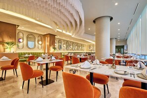 Uma sala de jantar ampla, com cadeiras laranja e mesas brancas, apresentando um design de teto moderno e grandes janelas.
