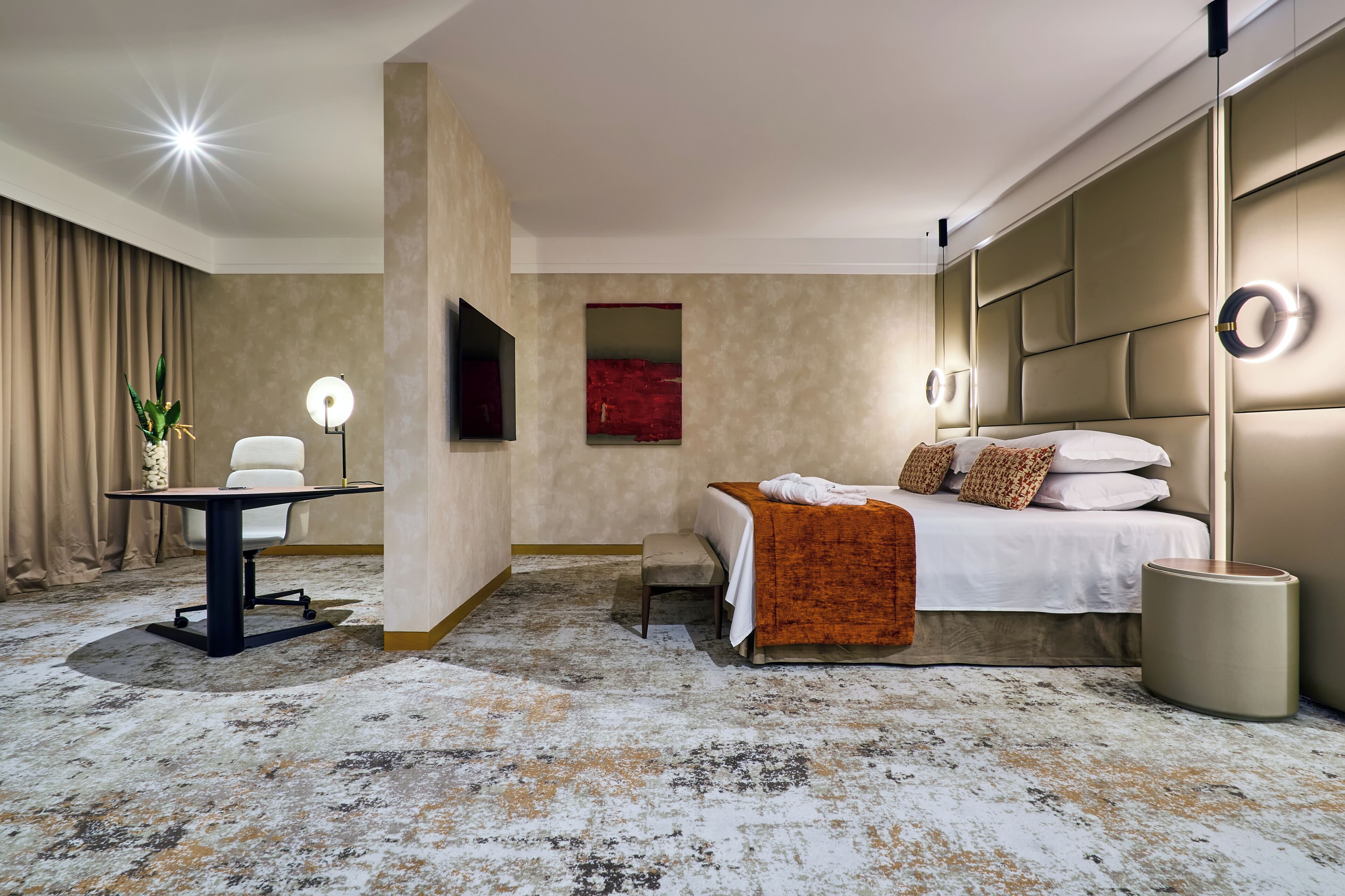 A hotel room with a bed, bedside table, chair, and a desk with a lamp.