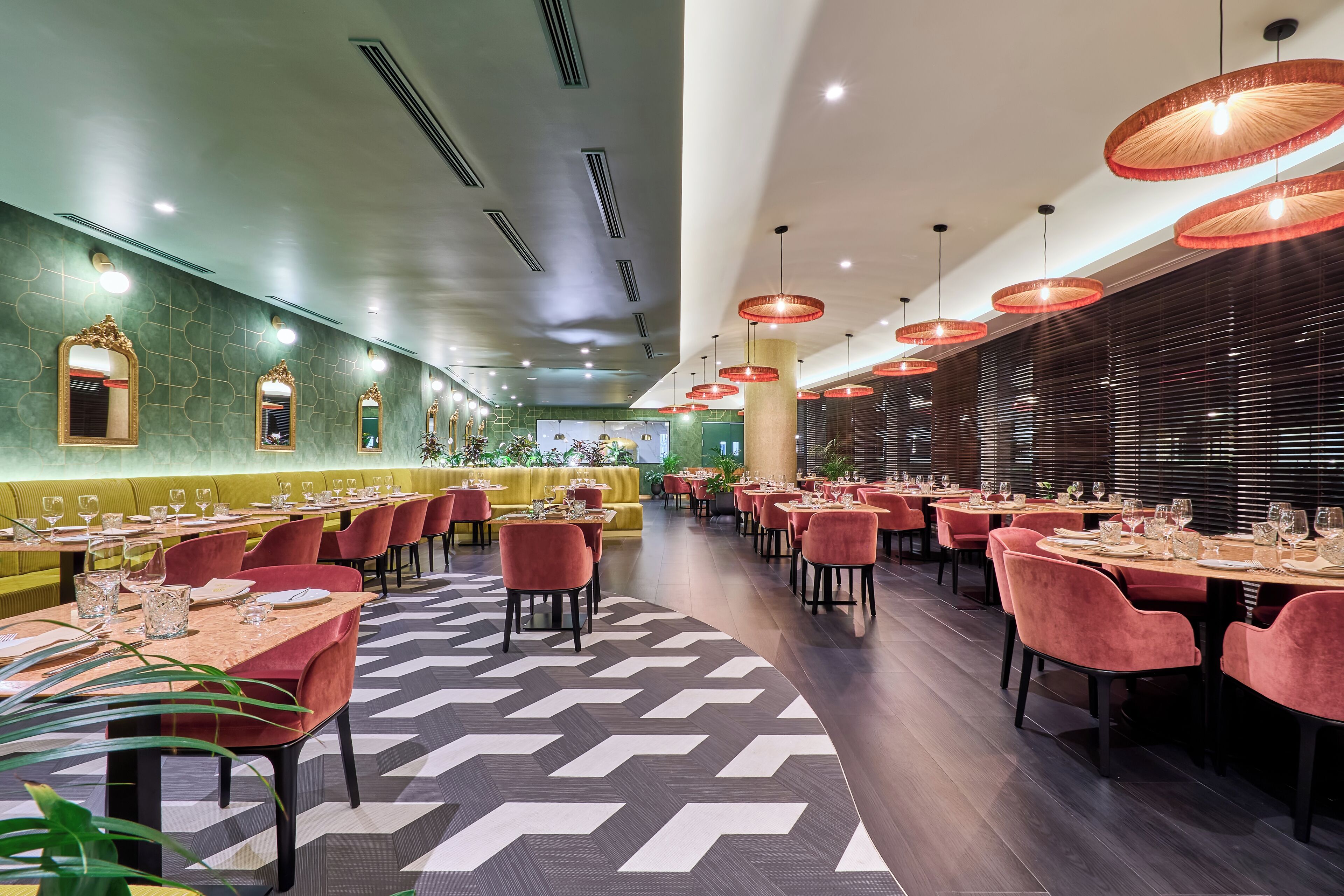 A restaurant with a modern interior, featuring a long dining area with tables and chairs, a patterned floor, and red pendant lights.