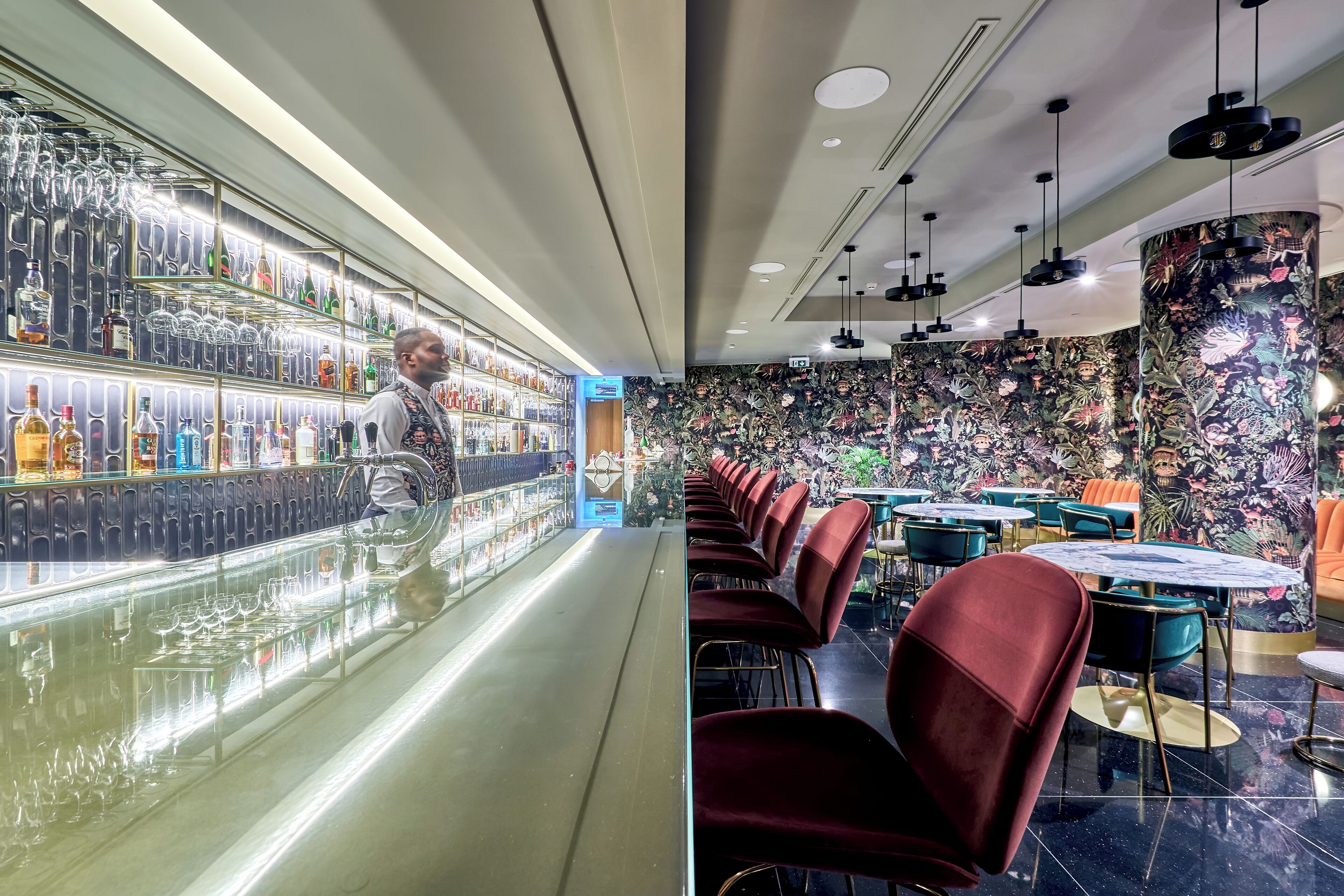 Zona de bar con estantes llenos de botellas y vasos, mostrador de bar y asientos con mesas redondas y sillas.