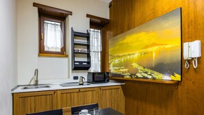 A modern kitchen with a wooden cabinet, a microwave, and a sink. There is a dining table with four cups and glasses. A painting of a bridge over water is on the wall.