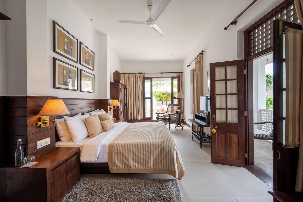 A spacious bedroom with a large bed, a desk, and a chair, a ceiling fan, and a view of the outdoors through a door.