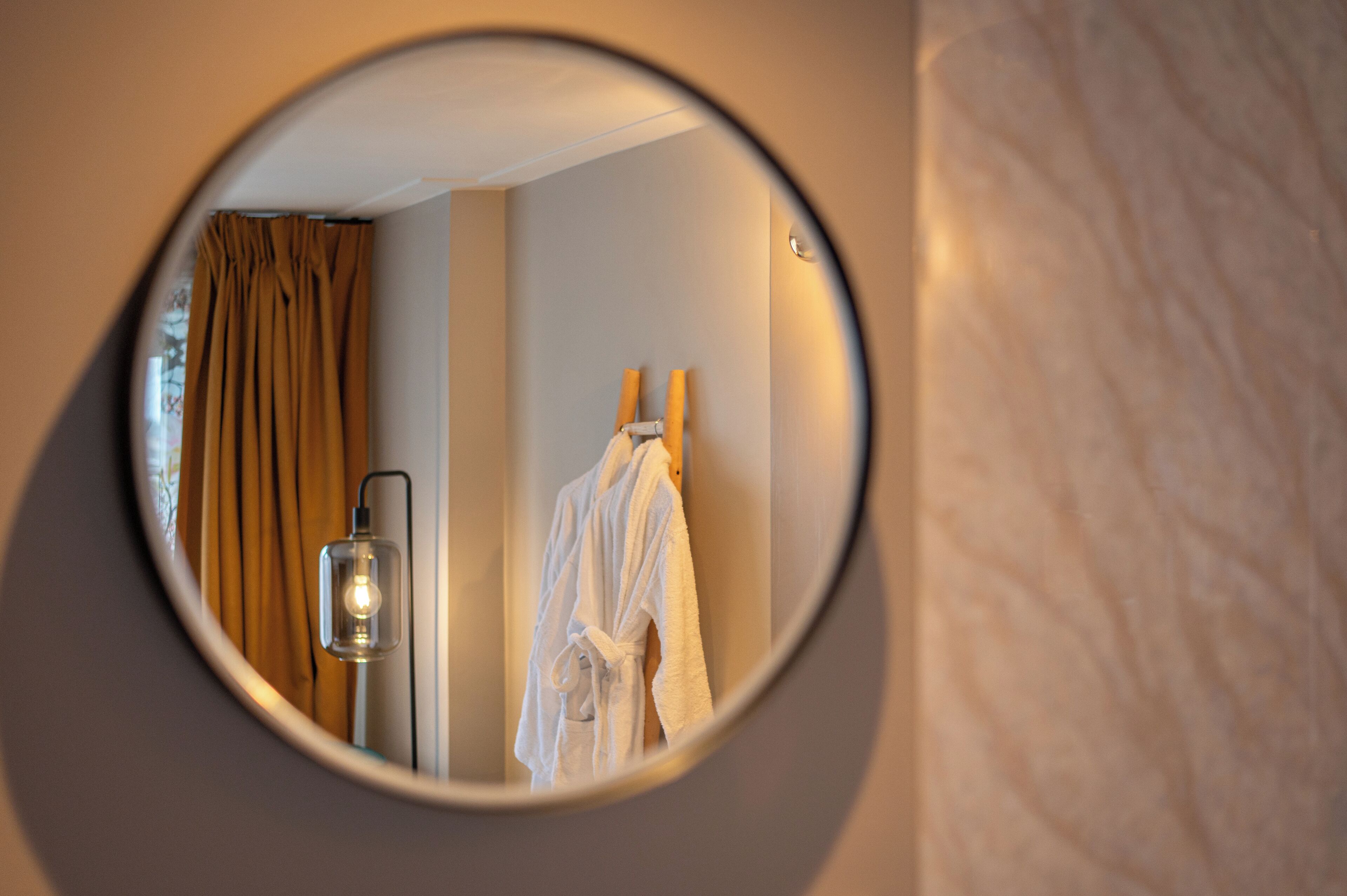 A round mirror reflecting a room with a white bathrobe hanging on a wooden hanger, a light fixture, and a brown curtain.