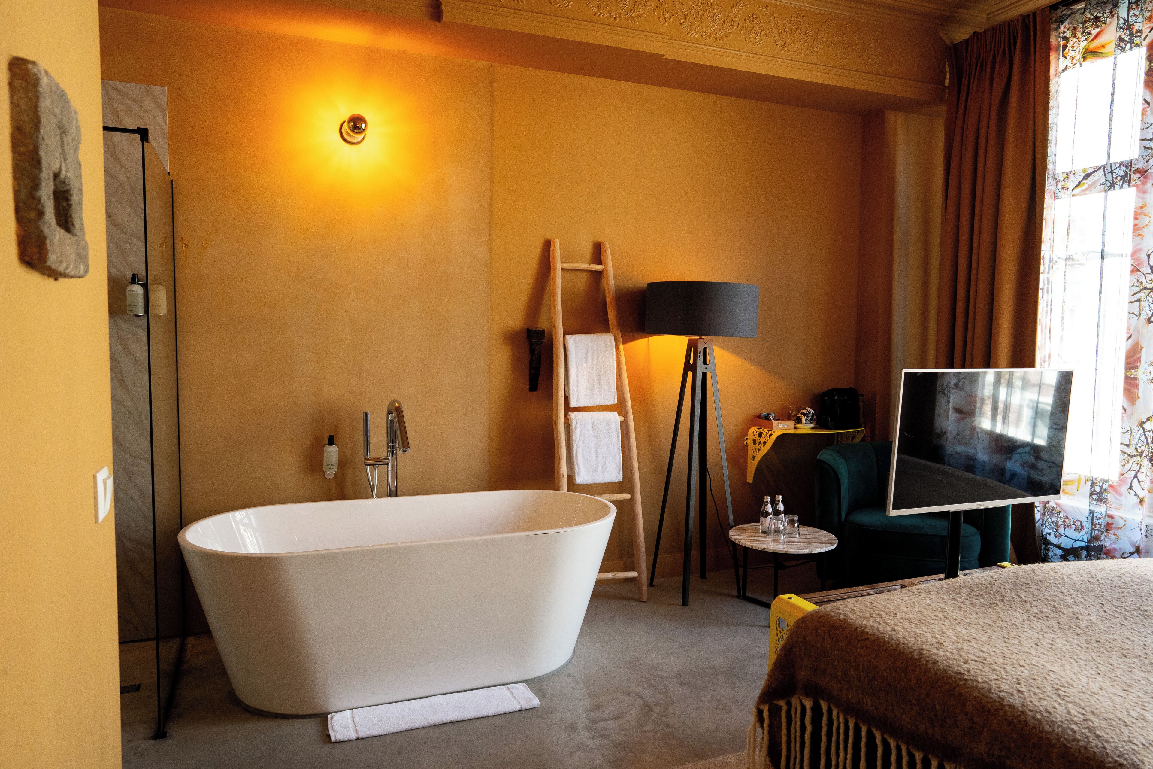 A modern bathroom with a freestanding bathtub, a towel rack, a small table with a lamp, and a television on a stand.
