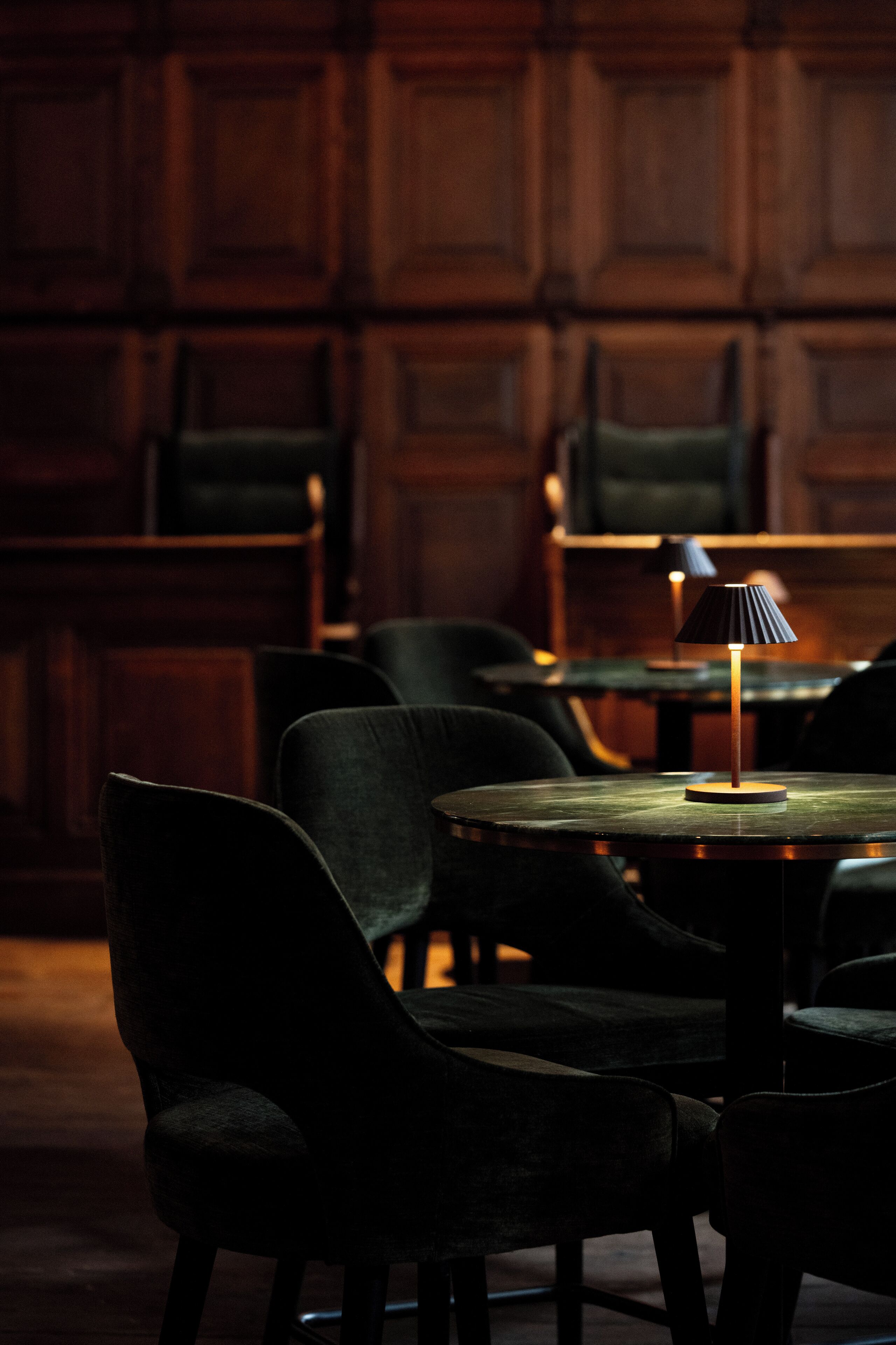 Een kamer met weinig licht, houten lambrisering, een ronde tafel met een lamp en meerdere donkere stoelen.