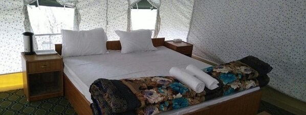 A neatly made bed with white linens and a floral-patterned blanket, a bedside table with a lamp, and a window with a view of trees. - The Ladakh Summer Camp  (Leh)