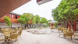 An outdoor seating area with tables and chairs, a central water feature, and surrounding trees.
