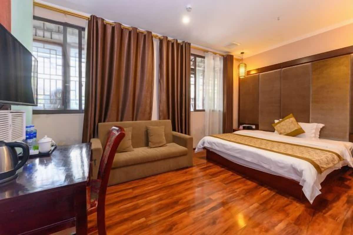 A hotel room with a wooden floor, a bed with white bedding and gold accents, a sofa, a desk with a kettle, and a window with curtains.