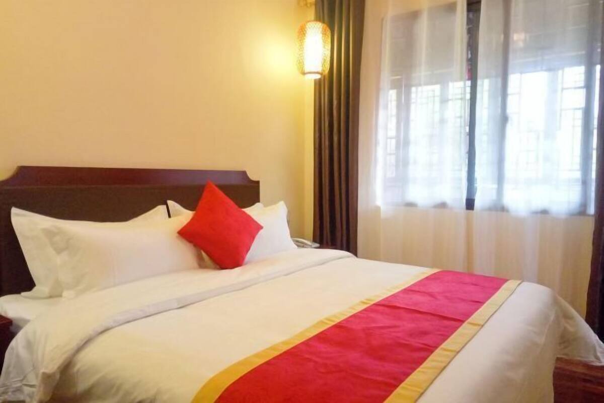 A hotel room with a bed, white bedding, a red and yellow bedspread, and a window with sheer curtains.